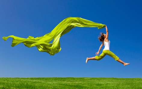 Beautiful young woman jumping on a green meadow with a colored tissue